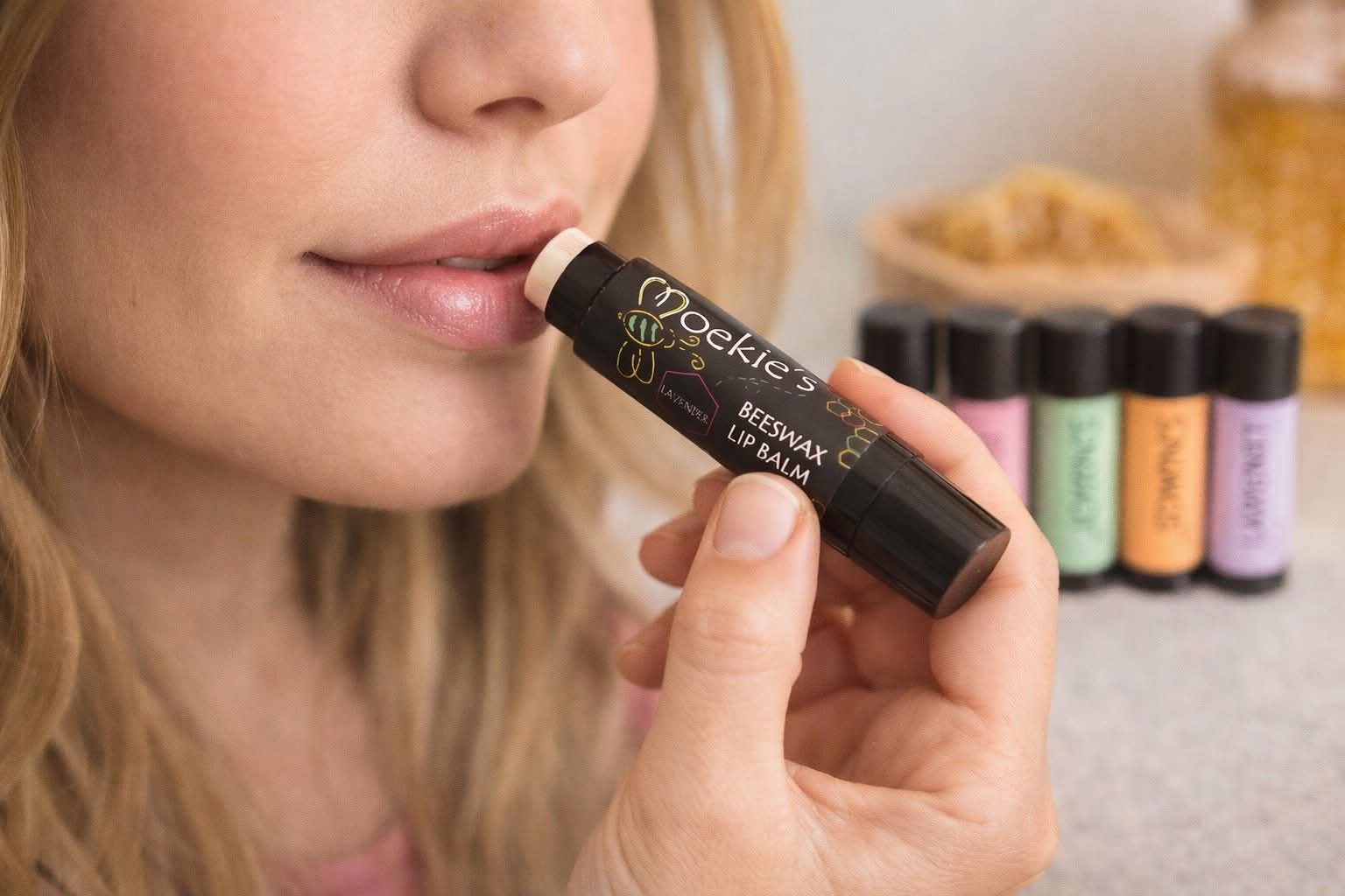 Woman applying Moekie's Beeswax Lip Balm in Lavender flavour, with multiple flavour tubes visible in the background